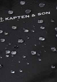 Svart texturerad tyg med ett hexagonalt mönster, som har flera vattendroppar och varumärket "KAPTEN & SON" tryckt i vitt.