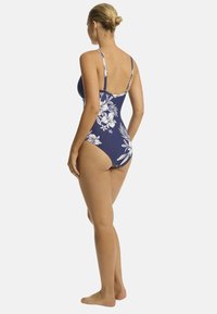 Een vrouw staat zijwaarts, gekleed in een marineblauwe badpak met witte bloemenpatronen, waarbij ze haar rug en benen toont tegen een effen achtergrond.