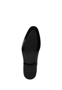 Suela de un zapato de vestir de cuero negro con área de agarre texturizada y tacón bajo apilado, vista desde abajo sobre un fondo blanco.