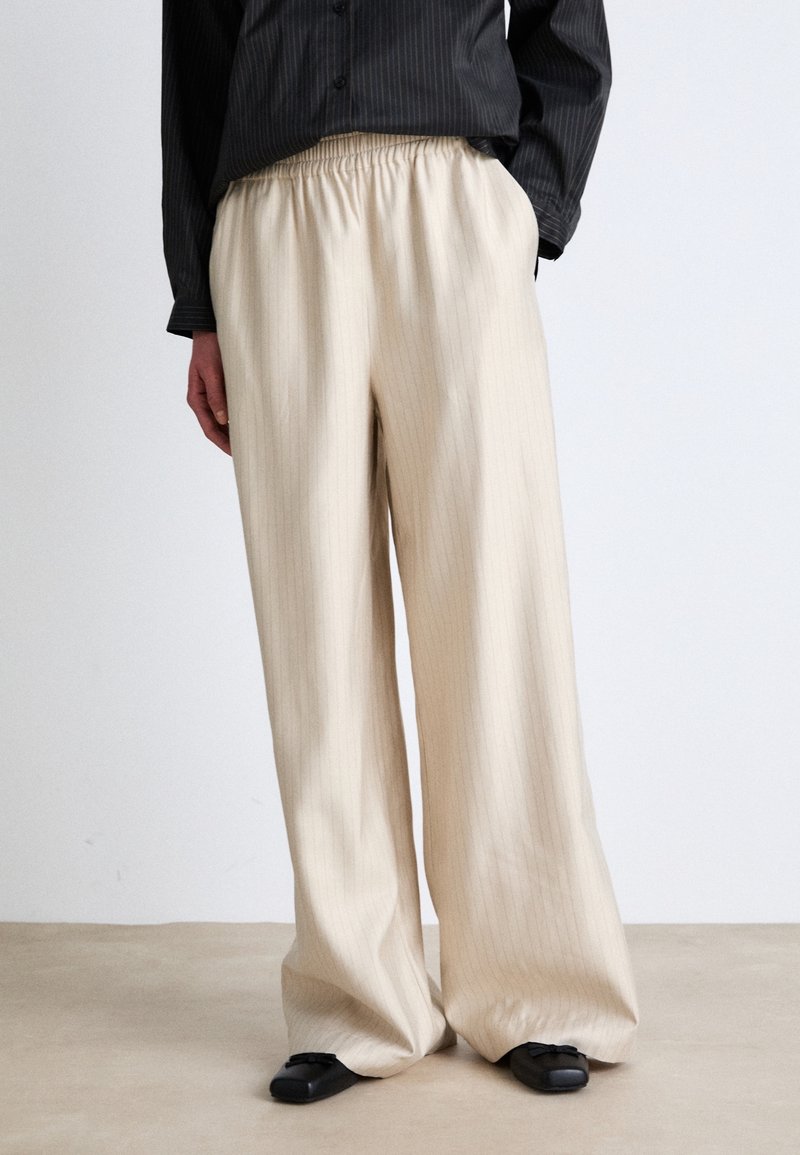 Pantalon beige ample à fines rayures avec taille élastique, porté avec des chaussures noires à bout carré et un haut noir à manches longues.