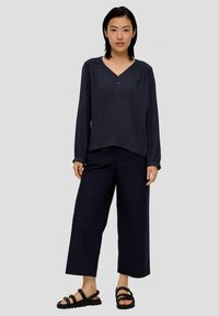 Navyblaues Langarmbluse mit V-Ausschnitt, Knopfdetails und strukturiertem Stoff. Kombiniert mit weit geschnittenen navyblauen Hosen und schwarzen Sandalen.