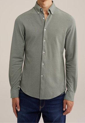 Homme portant une chemise boutonnée à manches longues, vert olive clair à col, avec un jean bleu foncé sur un fond neutre.