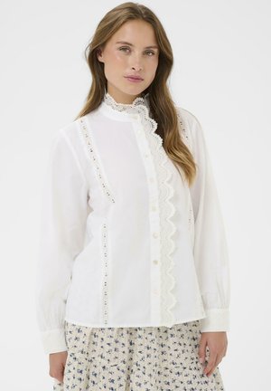 Frau, die eine weiße Bluse mit Spitzenbesatz und Perlenknöpfen sowie einen Rock mit Blumenmuster trägt, steht vor einem weißen Hintergrund.