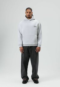 Lichtgrijze hoodie met buidelzak en "ZERO"-logo. Gecombineerd met losse zwarte spijkerbroek en zwarte schoenen met rode accenten. Gladde stoftextuur.