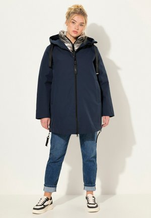 Marineblau wasserdichte Jacke mit Reißverschluss, Kapuze und einer inneren reflektierenden Schicht, kombiniert mit umgeschlagenen Jeans und schwarzen Turnschuhen.