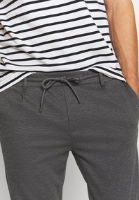 Homme portant un pantalon de survêtement gris foncé avec cordon de serrage et une chemise rayée horizontale blanche et noire, les mains dans les poches.