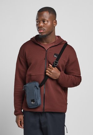 Haut de survêtement zippé matelassé marron avec capuche attachée. Un sac bandoulière bleu avec un logo et une sangle réglable, porté sur l'épaule.