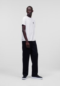 Witte T-shirt met korte mouwen en een kleine zwarte graphic op de borst, gecombineerd met losse zwarte broek en zwarte sneakers.
