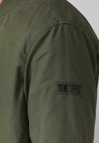 Olivgrüne Bomberjacke mit geripptem Kragen, glatter Stoffstruktur und einem schwarzen Patch mit dem Aufdruck "ELEVATED CASUAL EST. 1969" auf dem Ärmel.