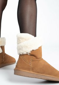 Botines de ante en tono arena con un puño de piel sintética blanca, suela plana de goma marrón y detalles de costura en el lateral; llevados con medias negras.