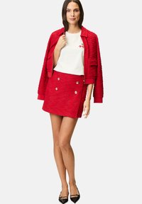 Red textured jacket with a matching mini skirt featuring gold buttons. White t-shirt displays a small cherry graphic. Black pointed toe flats.