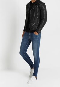 Hombre con jeans azules ajustados, chaqueta de cuero negra, camisa negra y zapatillas blancas, de pie con una mano en el bolsillo.