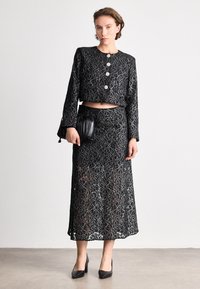 Juoda nėrinių dviejų dalių komplektas, kuriame yra trumpas švarkas su apvaliais mygtukais ir ilga sijonas. Papildytas maža juoda rankine ir aukštakulniais.