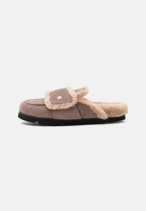 Chausson marron en daim à enfiler avec bordure et doublure en fourrure beige, semelle noire, et petit logo doré "SM" sur la languette supérieure.