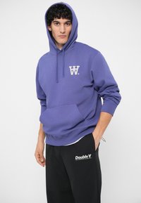 Fialová mikina s kapucňou, predným vreckom a logom. Mäkká látka, uvoľnený strih, rebrované manžety. Kombinovaná s čiernymi nohavicami s potlačeným logom.