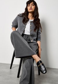Cardigan grigio a coste con bottoni dorati, t-shirt bianca, pantaloni a gamba larga grigio a righe e scarpe slip-on nere con accenti dorati.
