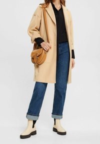 Cappotto oversize beige, camicia nera, jeans blu, stivali alla caviglia crema con accenti neri e borsa a tracolla marrone, con dettagli sottili.
