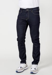 Dunkelblaue Denim-Jeans mit Slim-Fit, ausgestattet mit kontrastierenden gelben Nähten, fünf Taschen und einem Reißverschluss. Kombiniert mit weißen Turnschuhen.