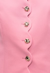 Ροζ ύφασμα με κυματιστή άκρη που έχει τέσσερα ακανόνιστα λαμπερά χρυσά κουμπιά ραμμένα κατά μήκος της άκρης.