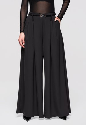 Žena nosící černé široké kalhoty s plisováním, tenký černý pás, průsvitný top s dlouhým rukávem, ruce v kapsách a černé špičaté boty.