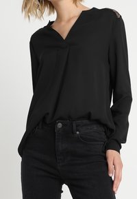 Blusa nera a maniche lunghe con scollo a V e dettagli in pizzo sulle spalle, abbinata a jeans scuri, creando una silhouette elegante.