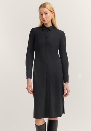 Eine Frau trägt ein schwarzes Midi-Kleid mit langen Ärmeln, Knopfleiste und Kragen, kombiniert mit schwarzen kniehohen Stiefeln, und steht vor einem schlichten Hintergrund.