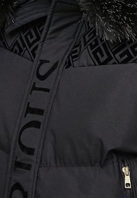 Doudoune noire avec col bordé de fourrure, doublure à motif géométrique, texte de marque vertical audacieux et poche zippée dorée.