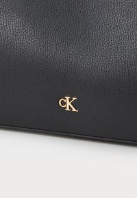 Juoda tekstūruota dirbtinė odinė rankinė su auksiniu "cK" logotipu. Turi elegantišką, stačiakampį dizainą ir lygų paviršių.
