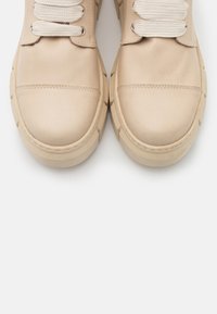 Baskets en cuir beige avec semelles en caoutchouc épaisses, dotées d'embouts ronds et de lacets en coton blanc. Texture lisse avec des détails de couture minimaux.