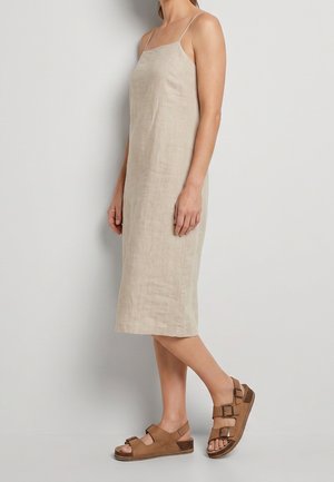 Kvinde iført en beige ærmeløs midi-kjole og brune sandaler med dobbelte spænder, som står mod en ensfarvet lys baggrund.