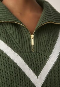 Detailní záběr na zelený pletený svetr s bílým žebrováním a zlatým zipem, který je č�ástečně rozepnutý u krku.