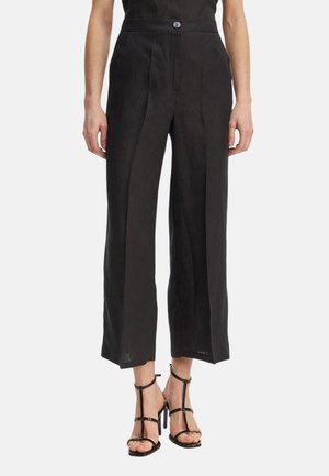 Donna che indossa pantaloni neri a vita alta, ampi e corti con pieghe anteriori e sandali neri con tacco alto e cinturini, su sfondo bianco.