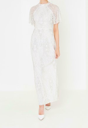 Kvinde iført en hvid gulvlængde kjole med korte gennemsigtige ærmer og indviklede sølvglimmermønstre, kombineret med hvide spidse hæle.