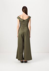 Olivegrønn jumpsuit med avskåret hals, vide ben og tettsittende bodice, med en diskret glidelås bak og minimale sømmer.