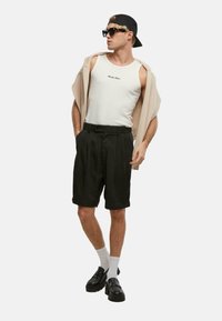 Hvit tanktopp med trykket tekst, beige genser drapert over skuldrene, svarte plissert shorts, svarte sko og hvite sokker. Tilbehør inkluderer solbriller og caps.
