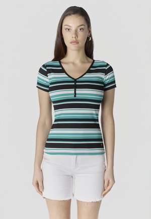 Joven mujer con una camiseta ajustada de rayas en tonos teal, negro y blanco con botones, y shorts de mezclilla blancos, de pie contra un fondo liso.