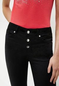 Jeans de denim negro con un diseño ajustado, que cuentan con cinco botones plateados en la cintura y una textura sutil. Combinados con una blusa roja con adornos.