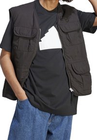 Czarna bezrękawnik z wieloma przednimi kieszeniami na czarnej koszulce z białym nadrukiem. Do tego luźne niebieskie dżinsy.