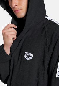 Sort frottébadekåbe med hætte, der har et hvidt broderet "arena" logo og mønstrede detaljer på ærmerne. Teksturen ser blød ud.