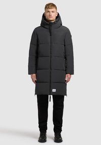 Dlhý čierny puffový kabát s kapucňou, vyrobený z hladkého materiálu. Má predný zips, bočné vrecká a značkovú etiketu na leme.