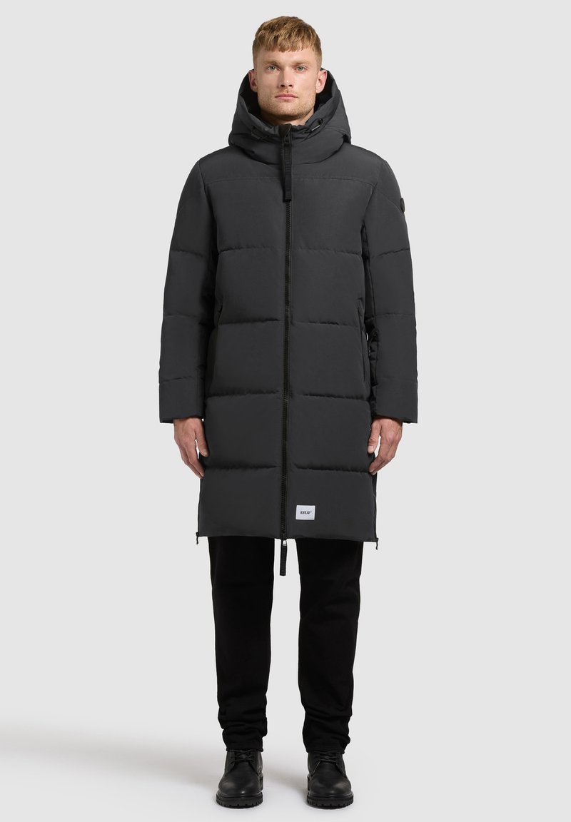 Długi czarny puchowy płaszcz z kapturem, wykonany z gładkiego materiału. Posiada zamek błyskawiczny z przodu, kieszenie boczne oraz metkę marki na dolnym brzegu.
