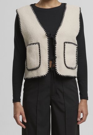 Personne portant une veste polaire crème avec surpiqûres noires et deux poches avant, par-dessus un haut noir à manches longues et un pantalon noir.