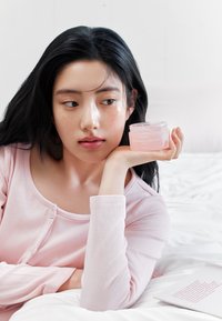 Barattolo rosa della maschera notturna al collagene Abib tenuto in mano, coperchio trasparente, consistenza liscia, posato su una coperta bianca. Tessuto rosa chiaro in alto.