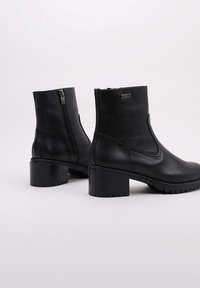 Botines de cuero negro con tacón grueso, cremallera lateral y textura suave; presentan un diseño minimalista y suela duradera.