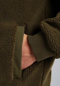 Zielona polarowa kurtka z teksturowanej tkaniny, z ręką w bocznej kieszeni i detalami na ściągaczach. Miękki, ciepły materiał.