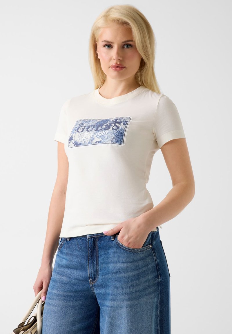 Femme aux cheveux blonds portant un t-shirt blanc avec le logo Guess 1981 et un jean bleu, tenant un sac à main beige avec une main dans la poche.