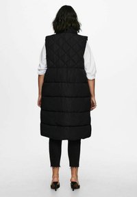 Juodas pūkinis liemenė su deimantiniu siuvinėjimu, be rankovių ir iki kelių, dėvima ant baltos marškinėlių, derinant su tamsiomis kelnėmis.