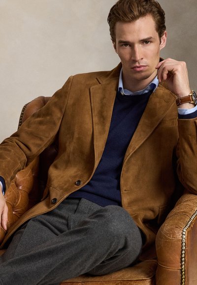 Mand sidder i en brun læderlænestol iført en brun ruskindsblazer, navy sweater, blå skjorte, grå bukser og et armbåndsur.