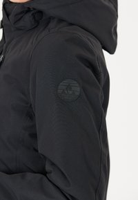 Schwarze wasserfeste Jacke mit Kapuze, aus strukturiertem Stoff und einem kreisförmigen schwarzen Logo auf dem linken Ärmel. Minimalistisches Design.