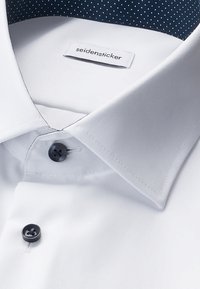 Weißes Hemd mit spitzem Kragen, zwei dunklen Knöpfen und einem sichtbaren "seidensticker"-Etikett im Inneren des Kragens vor einem dunkelblauen Hintergrund mit weißen Punkten.